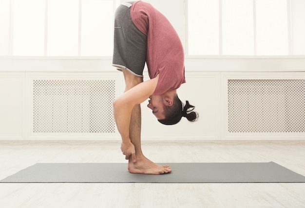 Hombre deportivo haciendo la postura de flexión hacia adelante de pie. Chico practicando yoga, párese de rodillas en el gimnasio, copie el espacio