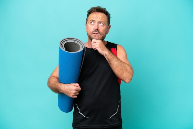 Hombre deportivo de mediana edad que va a clases de yoga mientras sostiene una alfombra aislada de fondo azul y mira hacia arriba