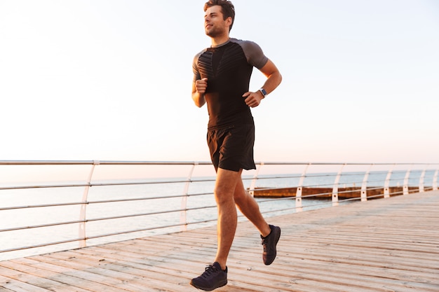 Hombre deportivo musculoso en pantalones cortos y camiseta corriendo a lo largo del muelle o paseo marítimo en la playa, durante el amanecer