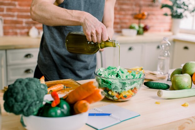 Un hombre deportivo prepara una ensalada en la cocina. Vierte aceite de oliva en una jarra de ensalada. Él está de pie en una cocina moderna en casa.