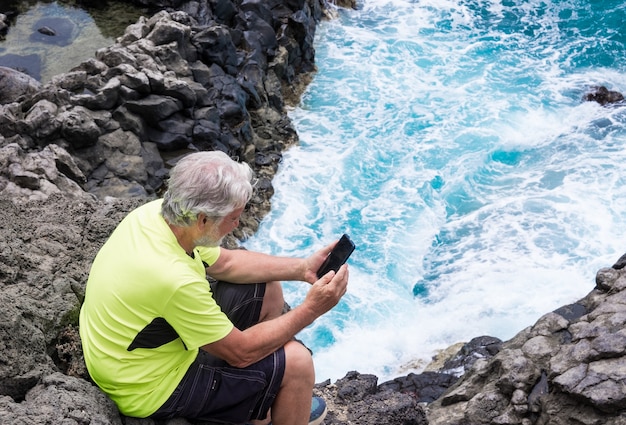 Hombre deportivo Senior en excursión al aire libre en el mar sentado en el acantilado mediante teléfono móvil. Cabello y barba blancos.