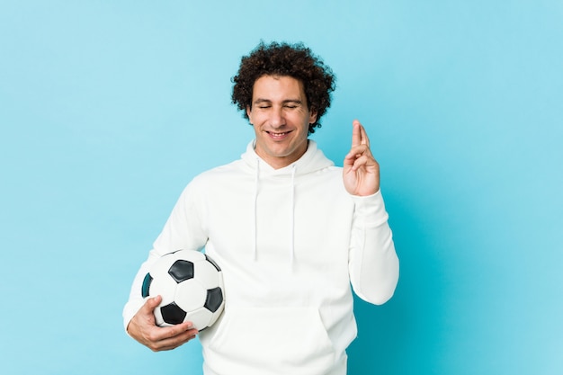 Hombre deportivo sosteniendo una pelota de fútbol cruzando los dedos para tener suerte