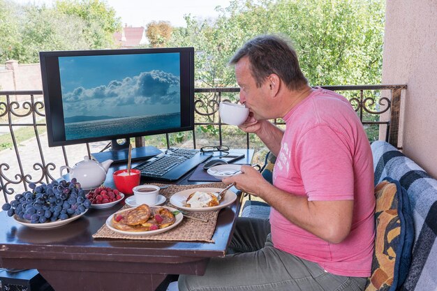 Hombre desayunando en el balcón de su casa cerca de la computadora