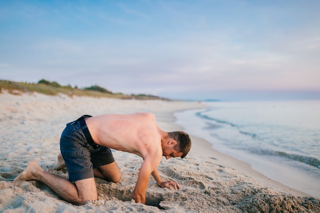 Hombre descalzo topless joven en pantalones cortos de pie sobre las rodillas y cavar hoyo con las manos en la playa más allá del mar en la noche de verano.