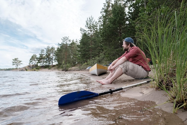 El hombre descansa en la orilla del lago después de viajar en kayak, se sienta tranquilamente en una playa de arena