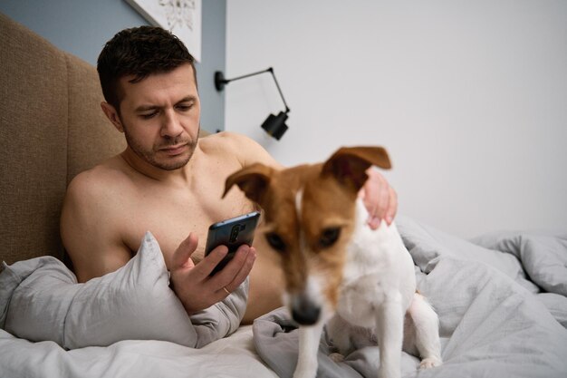 Hombre descansando en la cama con perro y usa smartphone