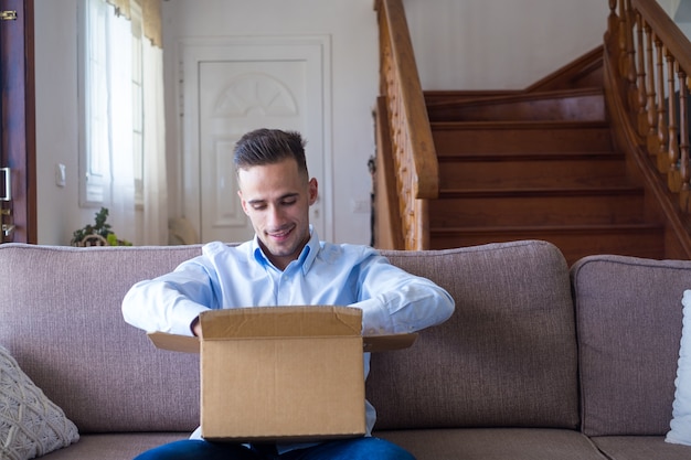 Hombre desembalaje del paquete de apertura de la caja de entrega en casa. Feliz joven mirando la caja de cartón mientras está sentado en el sofá en la sala de estar en casa. Hombre revisando cosas entregadas en casa.