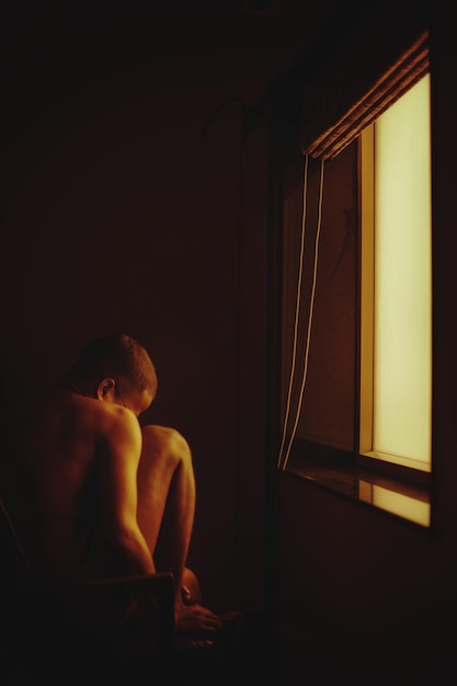 Hombre desnudo sentado junto a la ventana en casa