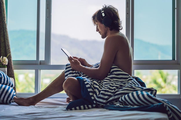 El hombre se despertó por la mañana y lee la noticia en la tableta en el fondo de una ventana con vista a las montañas.