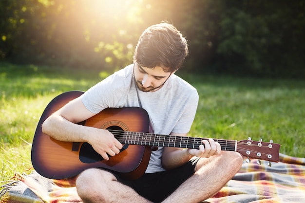 Hombre despreocupado con barba oscura y peinado moderno aprendiendo a tocar la guitarra mientras está sentado con las piernas cruzadas en la envoltura.