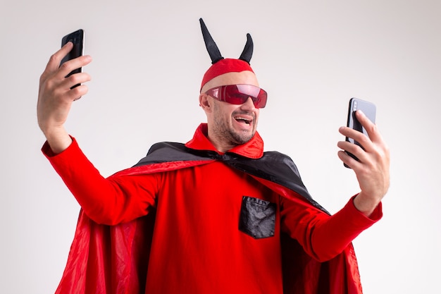 Hombre diabólico en traje rojo negro festivo de mascarada con pistola con dos teléfonos inteligentes en sus manos sobre estudio blanco.