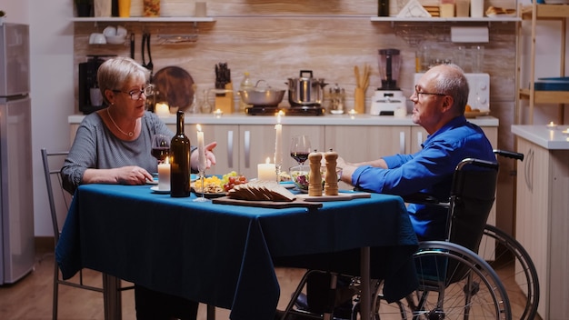 Hombre con discapacidad hablando con su esposa durante la cena festiva sentado a la mesa en la cocina. Marido en silla de ruedas inmovilizado discapacitado paralizado con celebración de aniversario.