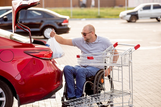 Foto hombre discapacitado adulto en silla de ruedas pone las compras en el maletero de un coche en el estacionamiento de un supermercado