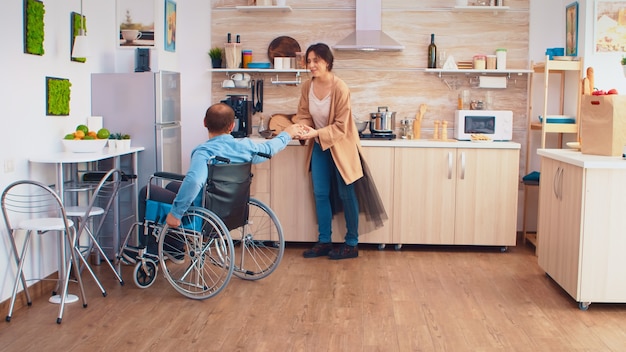 Hombre discapacitado en silla de ruedas abre el refrigerador mientras la esposa está cocinando y le da su caja de huevos. Chico con parálisis discapacidad discapacidad minusválidos dificultades para obtener ayuda para la movilidad de amor yr