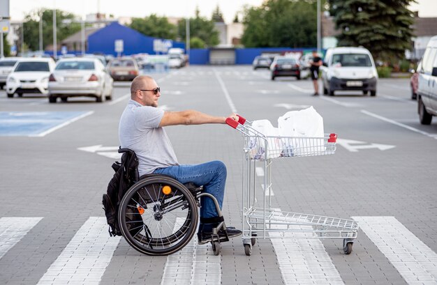 Hombre discapacitado en silla de ruedas empujando el carro delante de sí mismo en el estacionamiento del supermercado