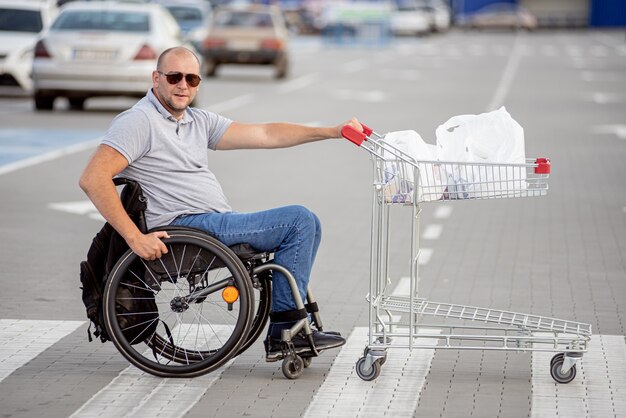 Hombre discapacitado en silla de ruedas empujando el carro delante de sí mismo en el estacionamiento del supermercado