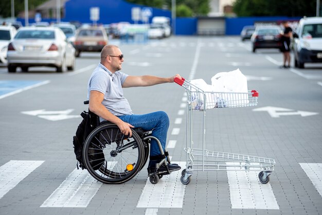 Hombre discapacitado en silla de ruedas empujando el carro delante de sí mismo en el estacionamiento del supermercado