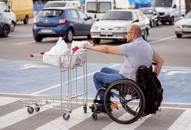 Hombre discapacitado en silla de ruedas empujando el carro delante de sí mismo en el estacionamiento del supermercado