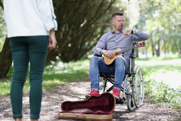 Hombre discapacitado en silla de ruedas sosteniendo ukelele y mirando su reloj mujer parada frente a