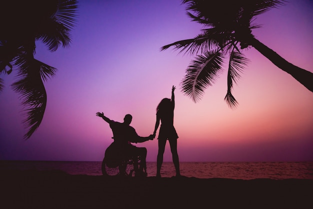 Hombre discapacitado en silla de ruedas con su esposa en la playa. Siluetas al atardecer