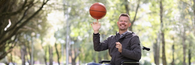 Hombre discapacitado sonriente gira la pelota de baloncesto en su dedo mientras está sentado en una silla de ruedas en el parque