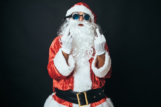 Hombre disfrazado de Papá Noel haciendo un gesto grosero, levanta los dedos en señal de 'vete a la mierda', sobre fondo negro. Concepto de Navidad, Santa Claus, regalos, celebración.