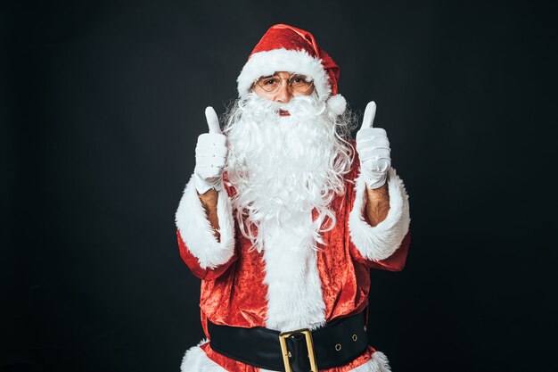 Hombre disfrazado de Papá Noel con los pulgares hacia arriba, sobre fondo negro. Concepto de Navidad, Santa Claus, regalos, celebración.