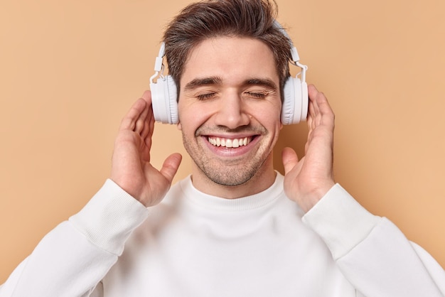 el hombre disfruta de unos auriculares estéreo con un sonido fantástico sonríe alegremente mantiene los ojos cerrados sonríe con dientes aislados en beige se siente vivo más que nunca.