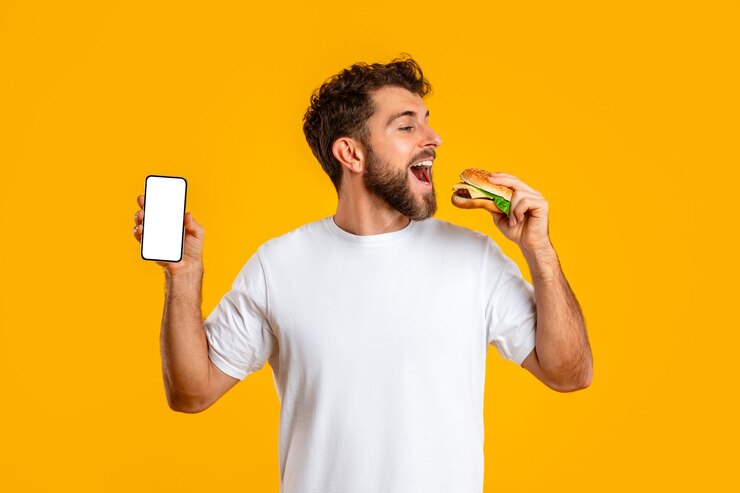 Hombre disfruta de una hamburguesa con queso mientras muestra un estudio de pantalla de teléfono móvil en blanco