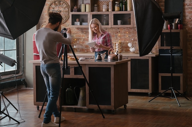 Hombre disparando blogger de comida. Sesión de fotos. Mujer en la encimera de la cocina con tableta. Fotografía entre bastidores.