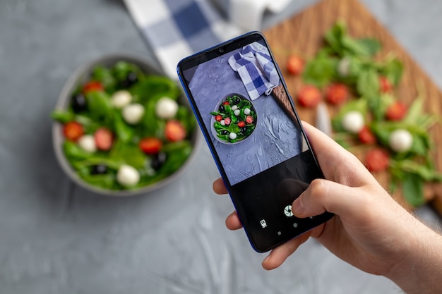 Hombre disparando ensalada de verduras frescas con mozzarella y espinacas en la cámara del teléfono celular. Cocinando