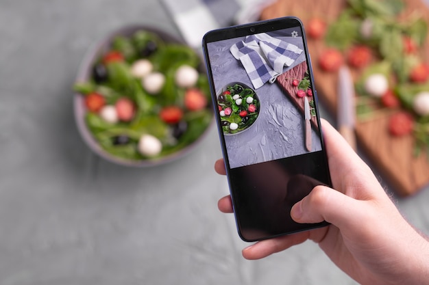 Hombre disparando ensalada de verduras frescas con mozzarella y espinacas en la cámara del teléfono celular. Concepto de cocina, blogs y alimentación saludable.