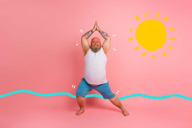 Hombre divertido con barba en posición de yoga sobre fondo rosa de estudio