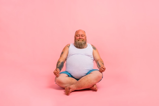 Hombre divertido con barba en posición de yoga sobre fondo rosa de estudio