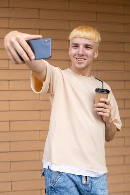 Hombre divertido vestido con una camiseta beige sosteniendo una taza de café en la mano y tomando selfies en un teléfono