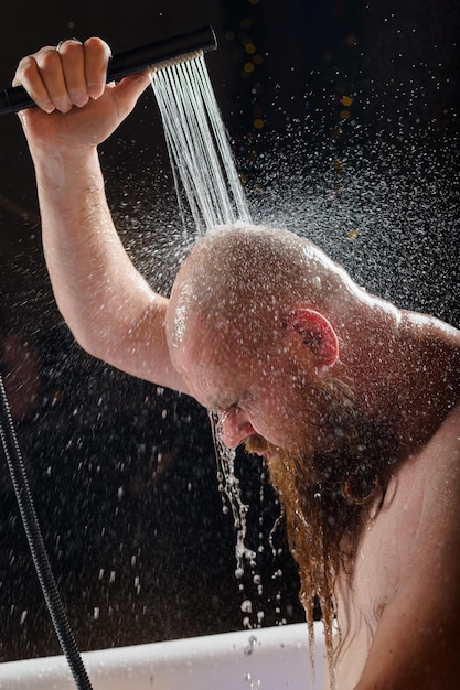 Foto el hombre se ducha un hombre brutal con barba roja está parado en el baño con agua corriente y lava la dispersión del aerosol sobre un fondo negro