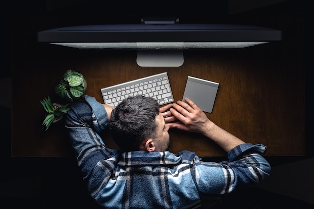 Un hombre duerme acostado frente a una computadora en su escritorio por la noche vista superior