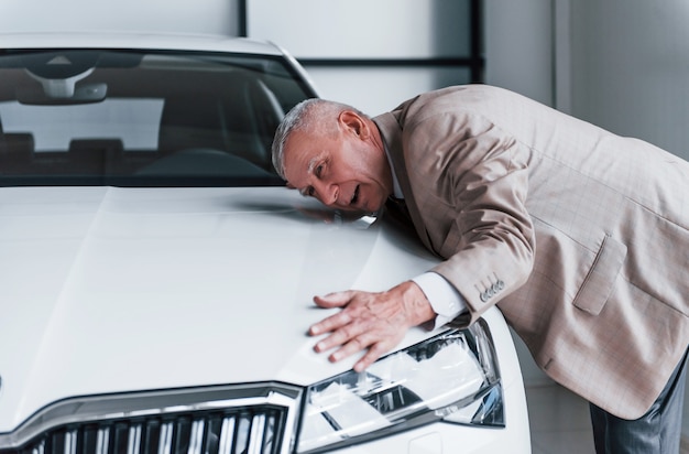 Hombre de edad alegre en ropa formal está parado frente al coche blanco moderno.