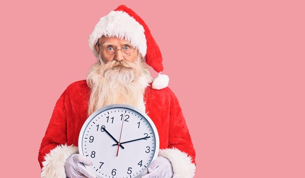 Hombre de edad avanzada con cabello gris y barba larga con disfraz de Santa Claus sosteniendo un reloj actitud de pensamiento y expresión sobria pareciendo seguro de sí mismo