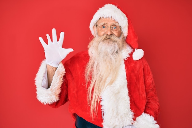 Hombre de edad avanzada con cabello gris y barba larga con traje tradicional de Santa Claus mostrando y señalando con los dedos número cinco mientras sonríe confiado y feliz