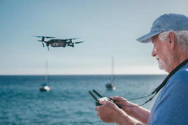 Hombre de edad avanzada volando con un dron al atardecer usando el control remoto Mar en el horizonte de fondo sobre el agua