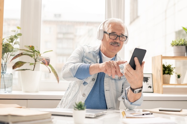Hombre de edad creativo en ropa casual y auriculares mirando a través de la lista de reproducción mientras está sentado en el lugar de trabajo en la oficina
