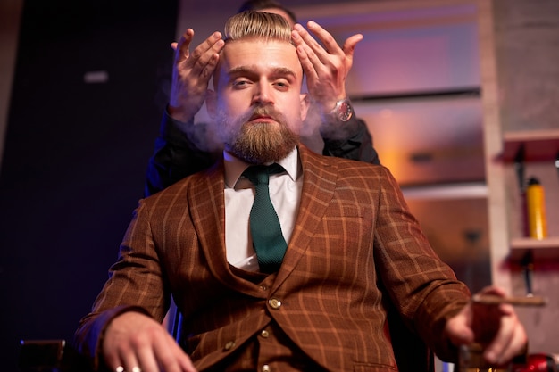 Hombre elegante disfruta de corte de pelo en la peluquería, fuma durante el proceso de corte