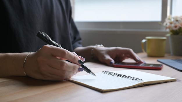 Hombre elegante escribiendo a mano en un cuaderno y usando un teléfono móvil para buscar información