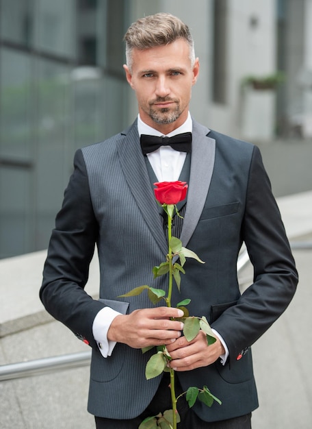 Hombre elegante en esmoquin negro hombre vestido con esmoquin corbatín al aire libre grizzle esmoquin hombre con rosa roja