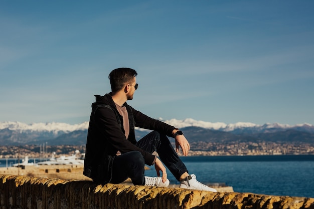 Hombre elegante con gafas disfrutando de la vista del mar y la montaña nevada.