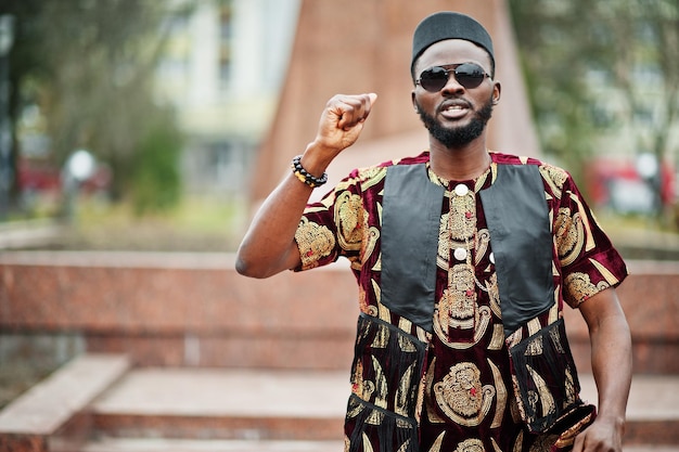 Hombre elegante y guapo africano con traje tradicional y gorra negra levantó su mano derecha contra el monumento.