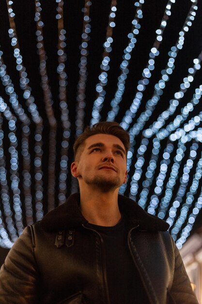 Hombre elegante y guapo de moda con el pelo vestido con ropa de moda con una chaqueta de cuero negro sobre un fondo de luces azules en la ciudad Hombre profesional y tecnología