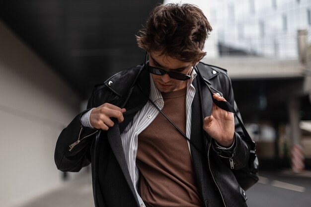 Foto hombre elegante joven inconformista fresco en gafas de sol oscuras de moda en una camisa con una chaqueta de moda vintage posando en la ciudad en un día de primavera. chico atractivo en ropa juvenil en la calle. estilo americano moderno