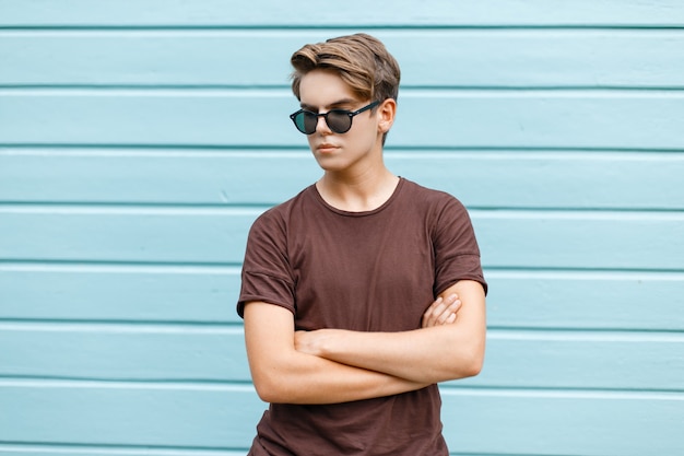 Hombre elegante joven inconformista con un peinado de moda en gafas de sol oscuras en una camiseta marrón de moda posando cerca de una pared de madera azul en un día cálido y soleado de verano